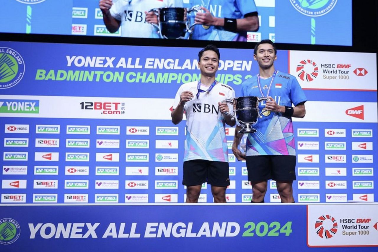 Momen Jonatan Christie mengajak Anthony Ginting memegang trofi All England 2024 sektor tunggal putra di podium All England, Utilita Arena Birmingham, Minggu (17/3/2024).